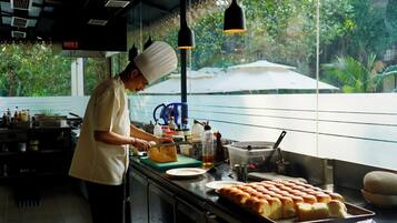 A kitchen with a chef preparing food, a large window with a view of greenery, and a tray of baked goods.