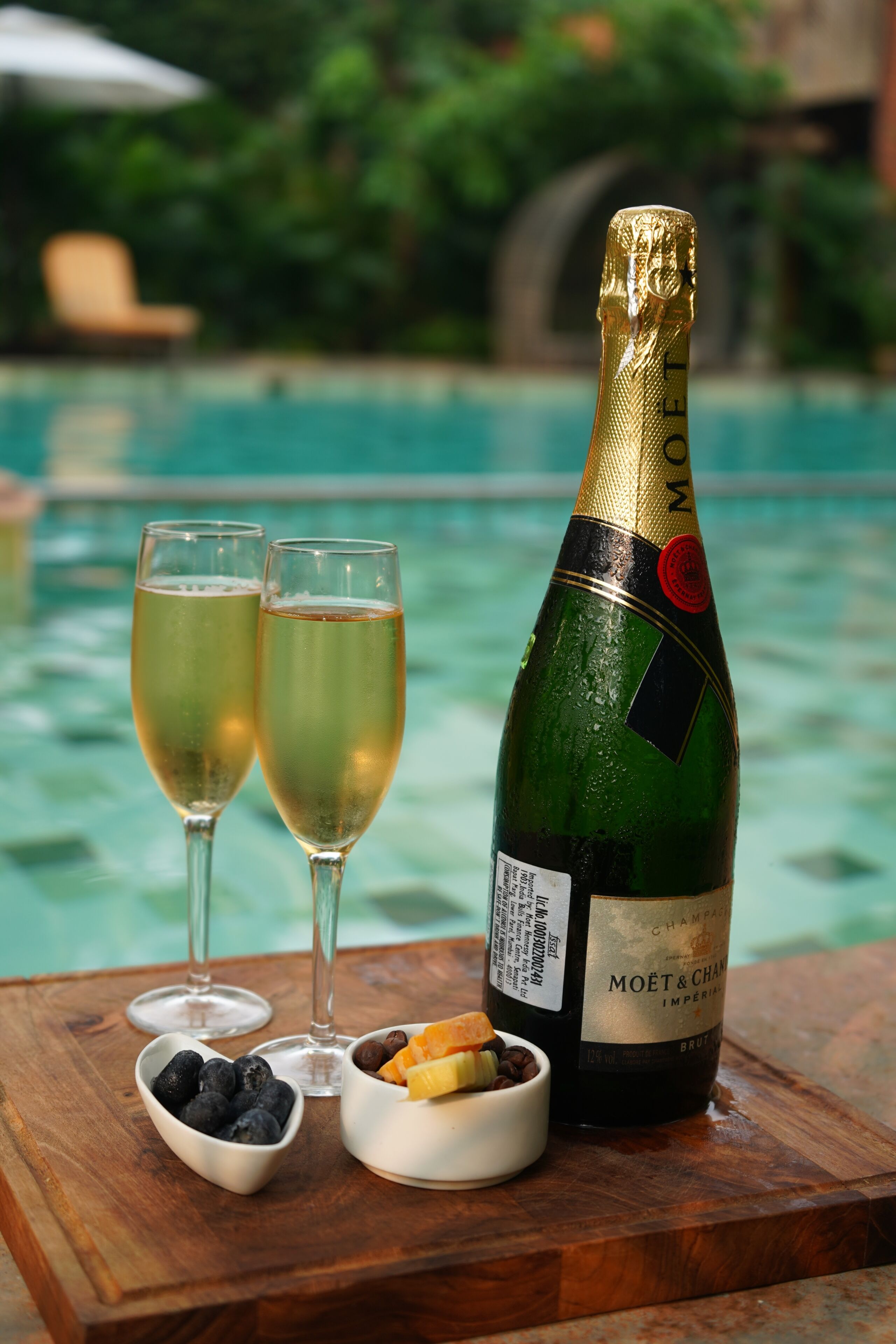 A bottle of Moët & Chandon champagne, two glasses, and small bowls of fruit and coffee on a wooden table by a pool.