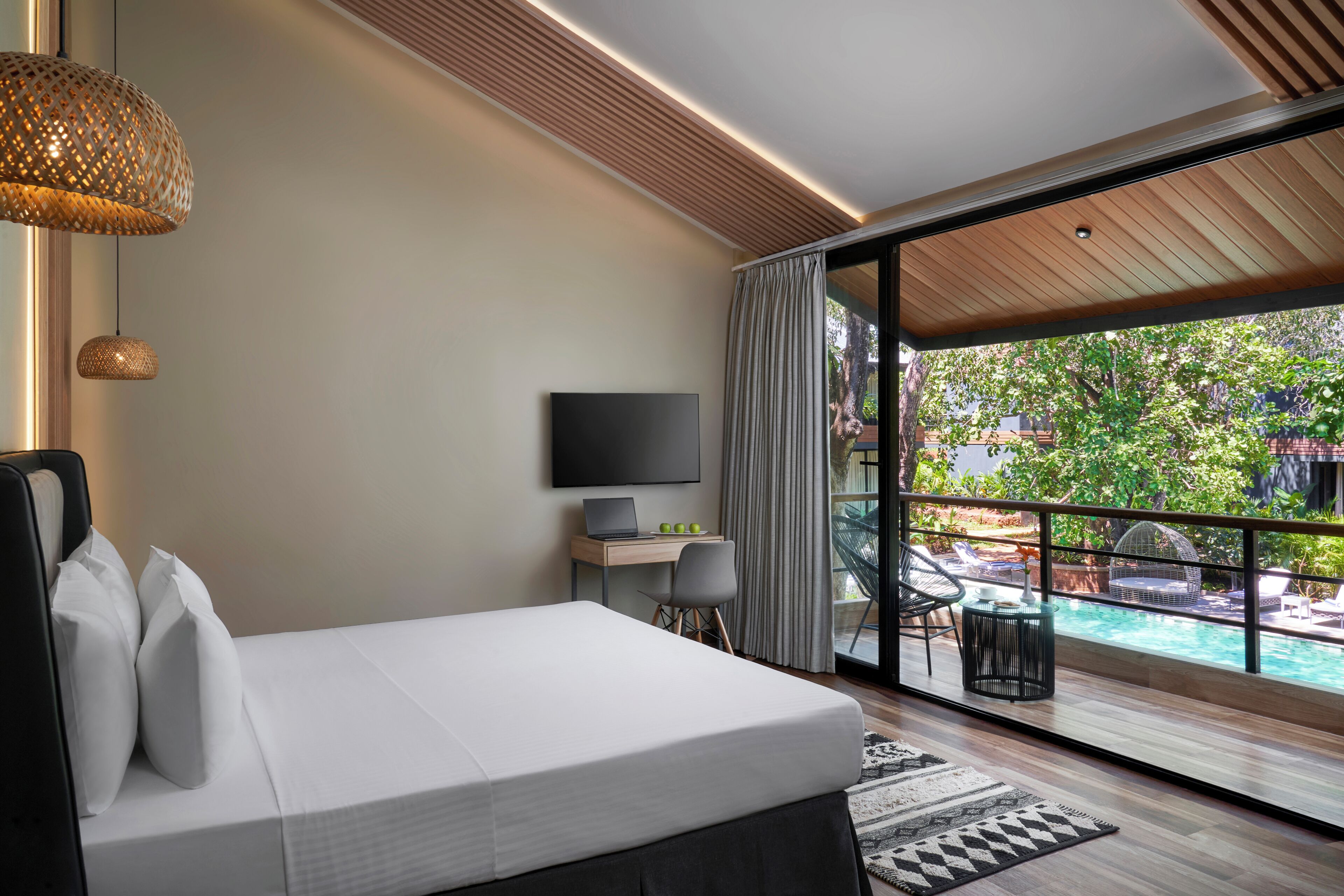 A modern hotel room with a bed, a desk with a laptop, a TV, and a view of a swimming pool.