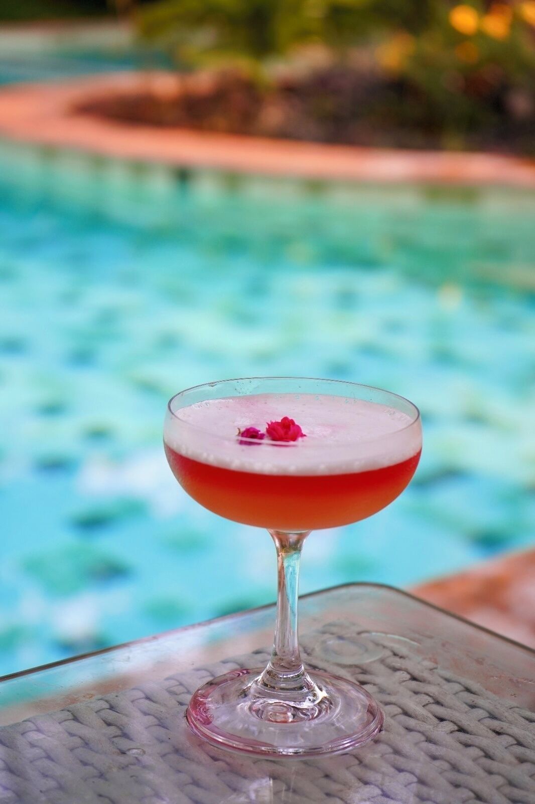 Un vaso de cóctel con una bebida roja y una decoración floral, sobre una mesa junto a una piscina.