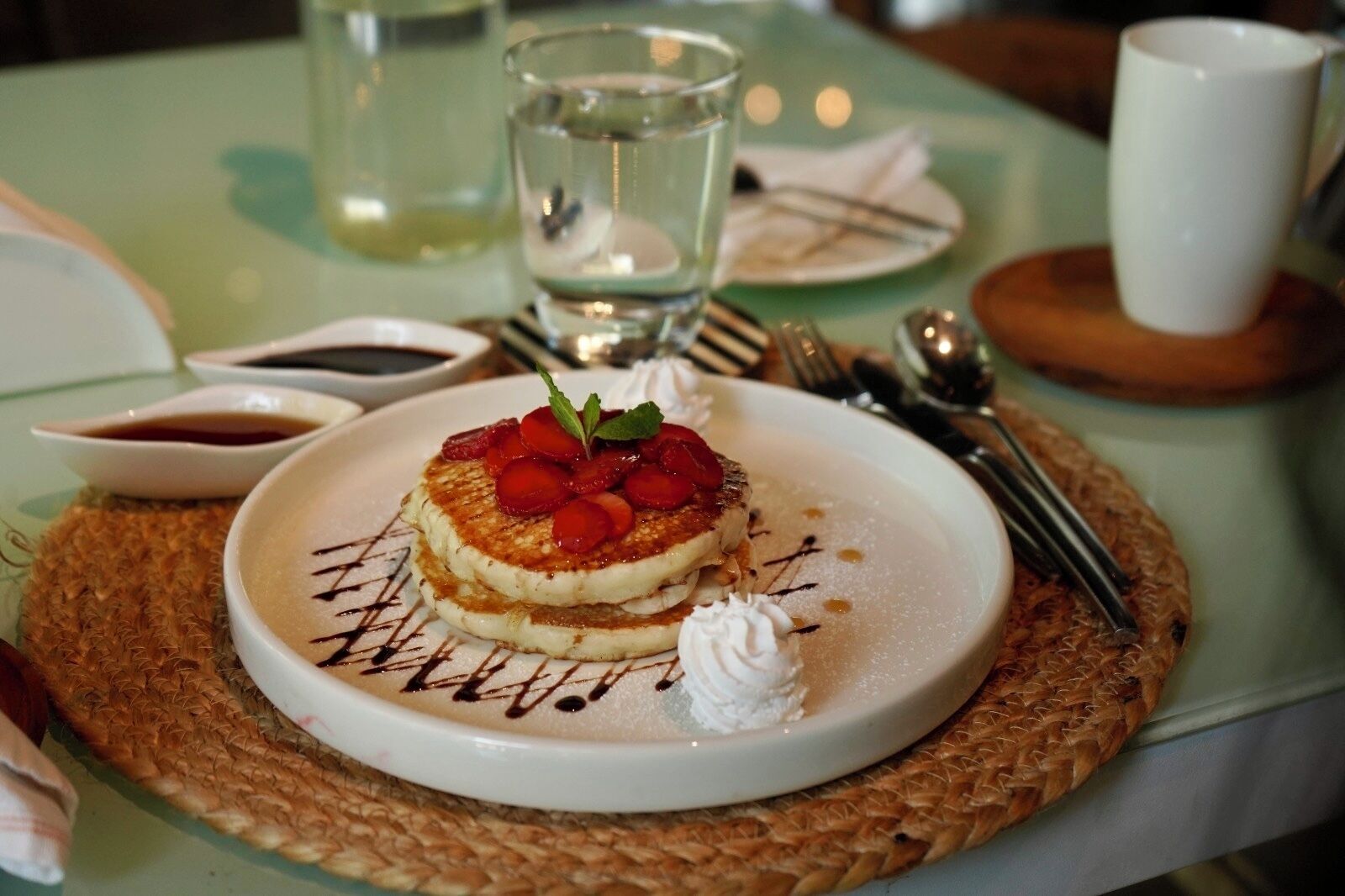 Un plato de postre con un panqueque, fresas, crema batida y un chorrito de chocolate sobre un posavasos tejido.