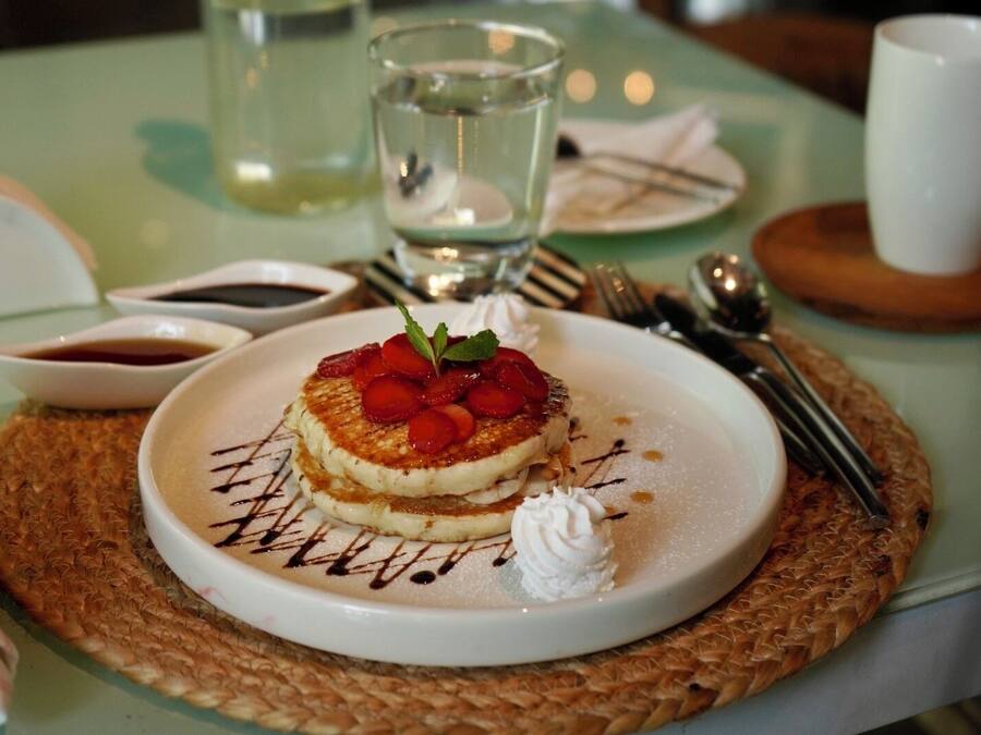 A dessert plate with a pancake, strawberries, whipped cream, and chocolate drizzle on a woven placemat.