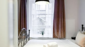 Un lit bien fait, avec une couette blanche et une serviette pliée, un luminaire suspendu noir et une fenêtre avec des rideaux.