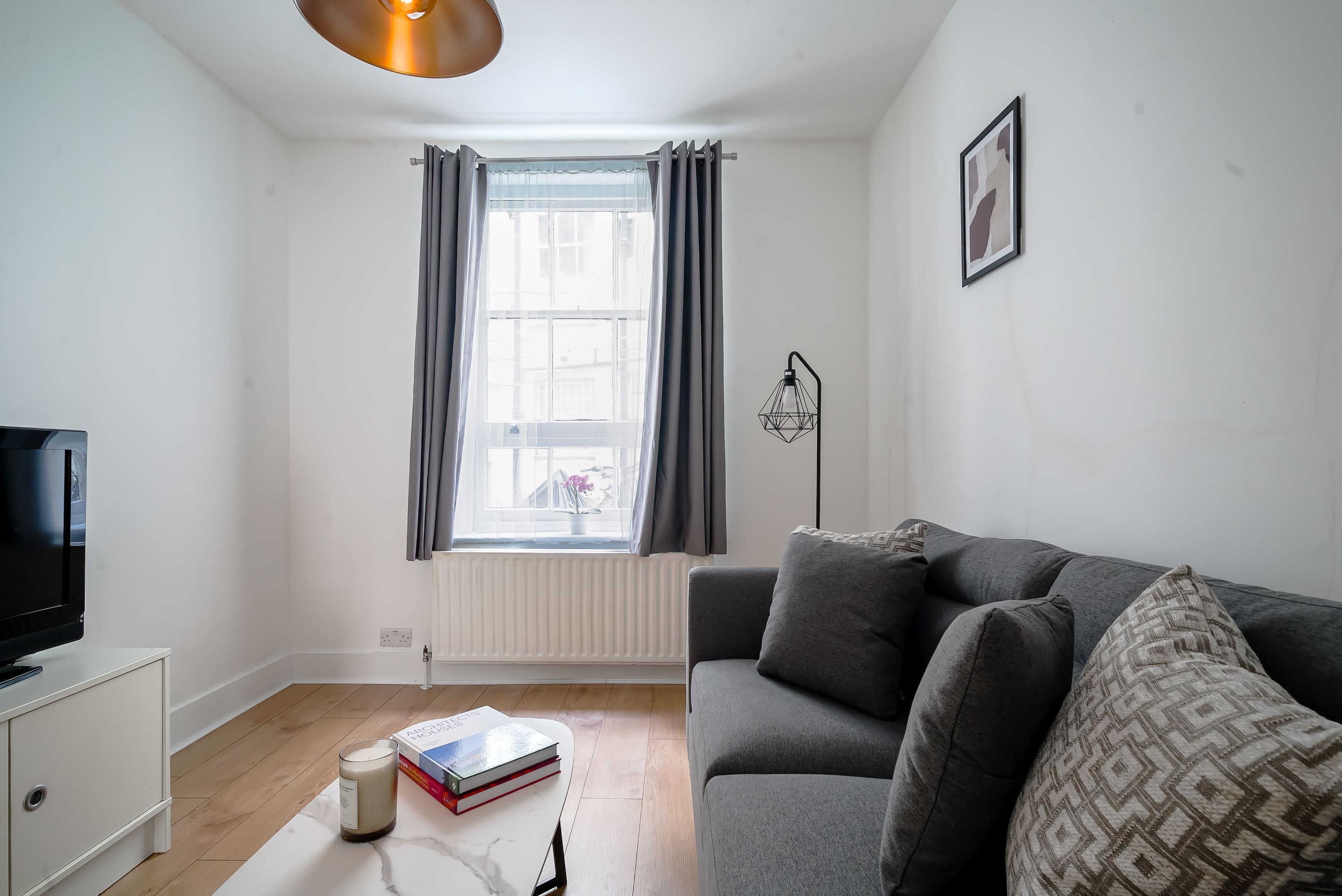 A modern living room with a grey sofa, a white coffee table, a book, a candle, and a window with grey curtains.