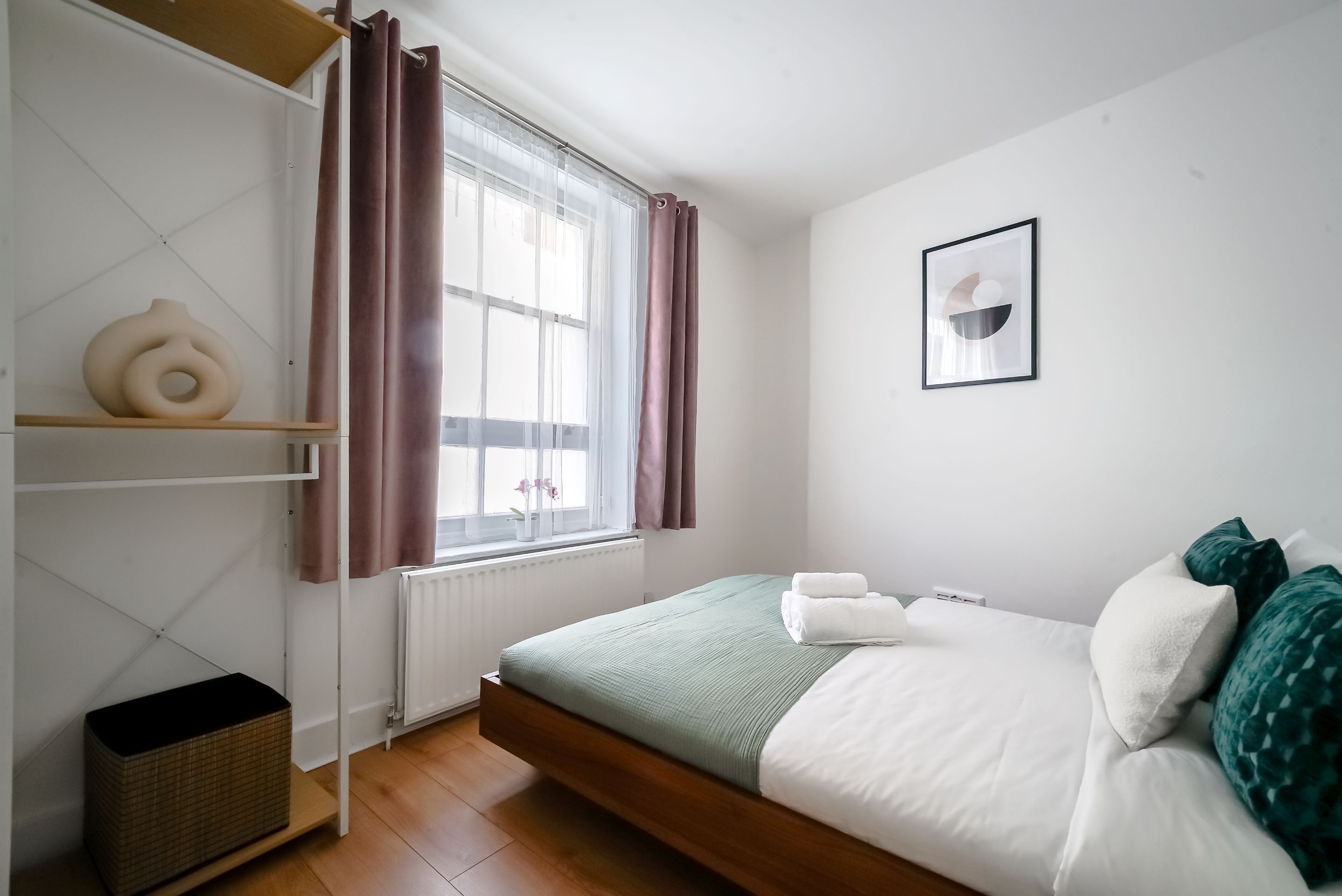 A minimalist bedroom with a bed, a window with curtains, a radiator, and a wall-mounted picture.