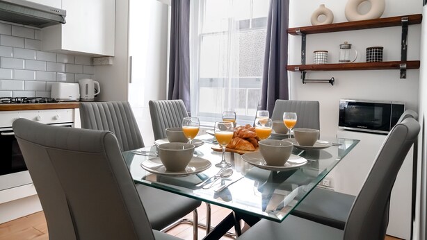 Une cuisine moderne avec des meubles blancs, une table à manger en verre et un coin petit-déjeuner avec des croissants et du jus d’orange.