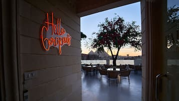 A hotel entrance with a neon sign reading "Hello Guests," a view of the pyramids, and outdoor seating area.