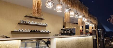 A bar area with a thatched roof, hanging lights, and shelves with glasses and bottles.