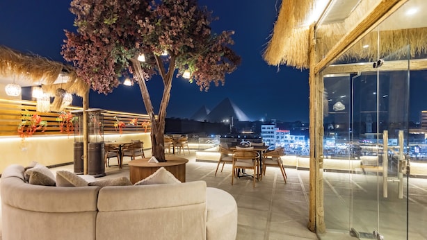 Un lounge en la azotea con zona de estar acolchada, un árbol y pared de vidrio con vista a la ciudad.