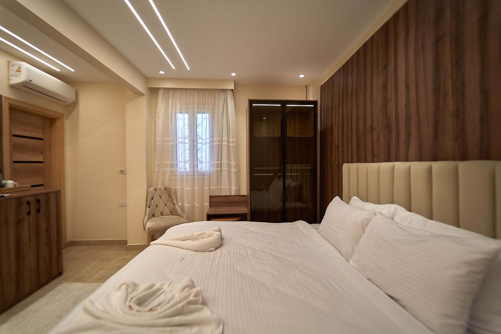 Un dormitorio moderno con una cama grande, un armario de madera, una silla y un armario con puerta corredera de vidrio.