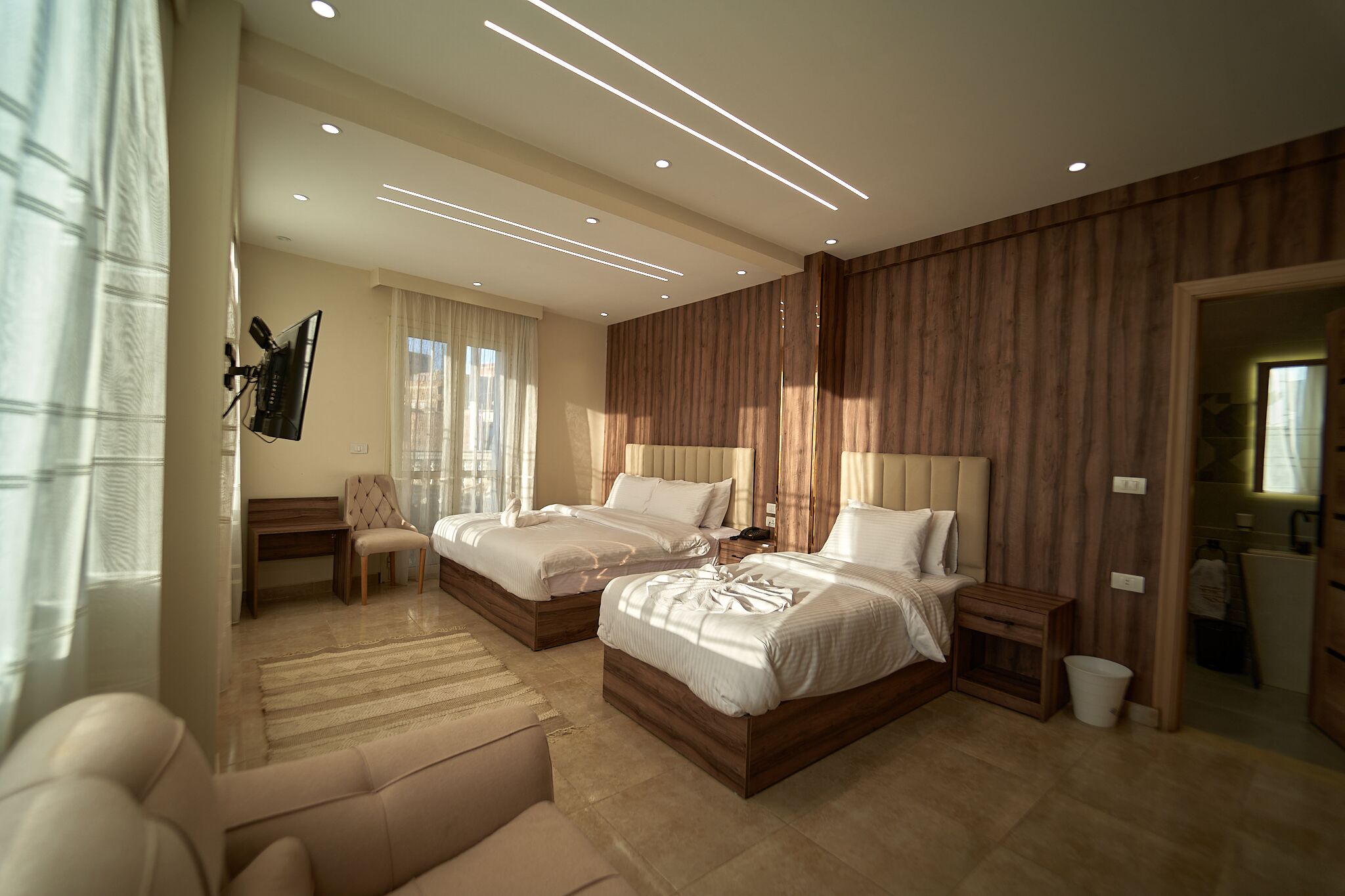 A modern hotel room with two beds, a sofa, a desk, and a bathroom visible through an open door.