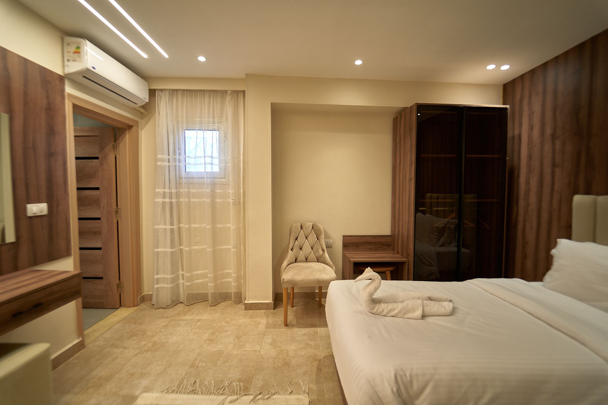 A hotel room with a bed, a chair, a desk, an air conditioning unit, a wardrobe, and a window with curtains.