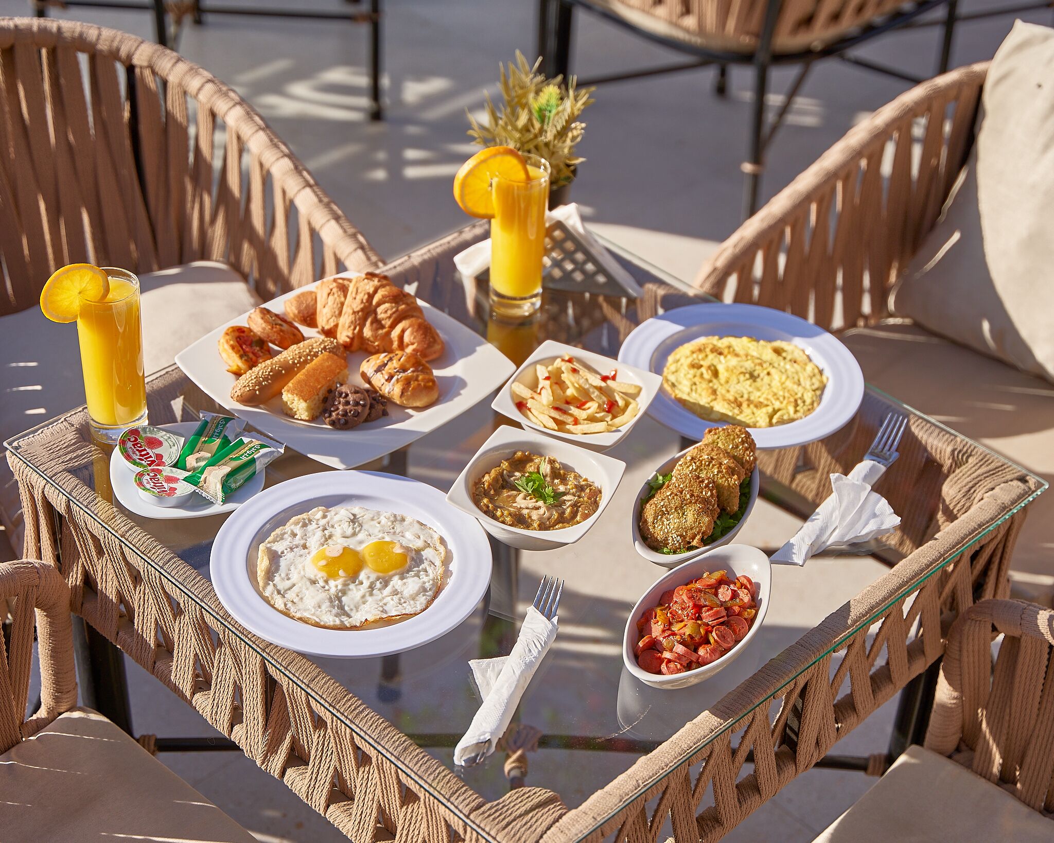 A breakfast tray with croissants, eggs, sausages, and a glass of orange juice on a wicker table outdoors.
