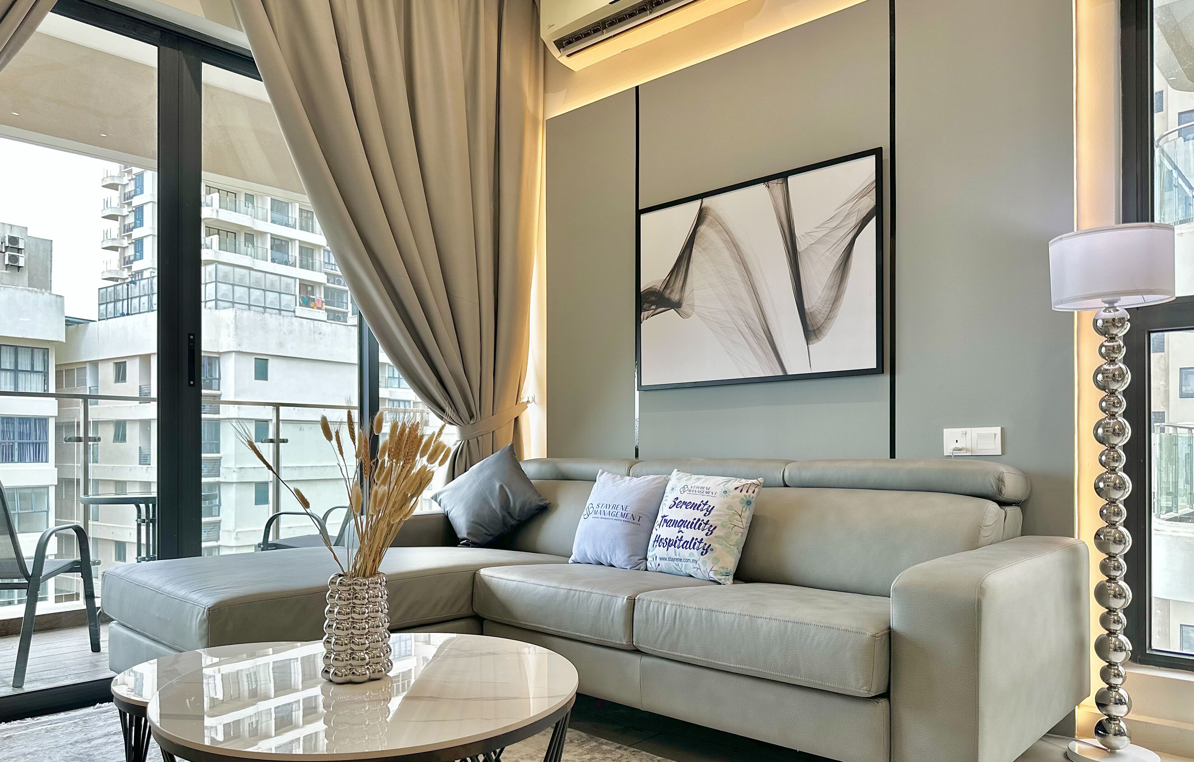 A modern living room with a sofa, a round coffee table, and a vase with dried plants. There is a large window with curtains, a wall-mounted air conditioner, and a framed abstract painting.