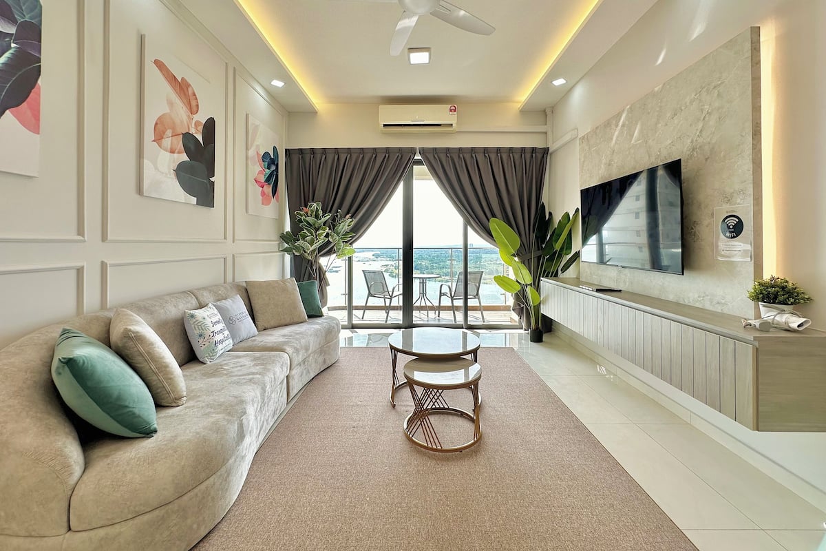 A modern living room with a sofa, a coffee table, and a TV. There is a balcony with a view.