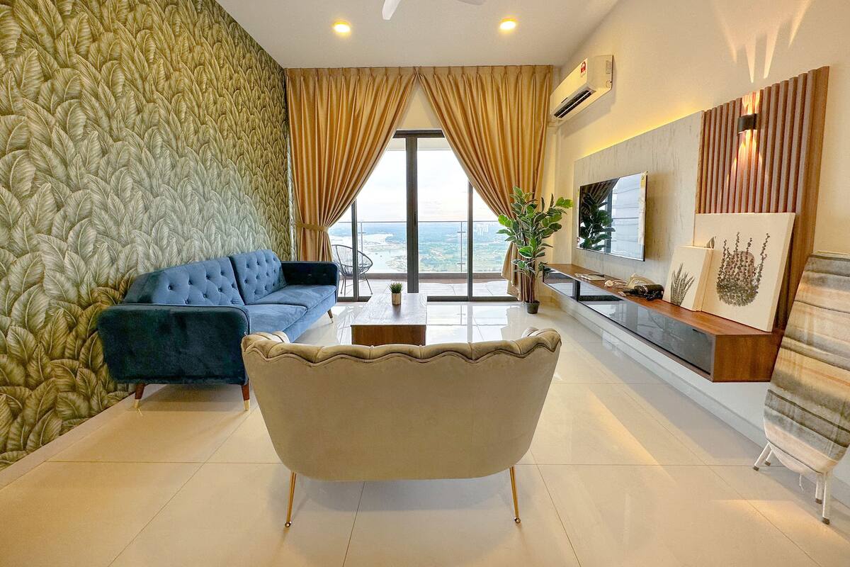 A modern living room with a blue sofa, a beige armchair, and a glass coffee table. There is a large window with curtains, a ceiling fan, and a flat-screen TV mounted on the wall.
