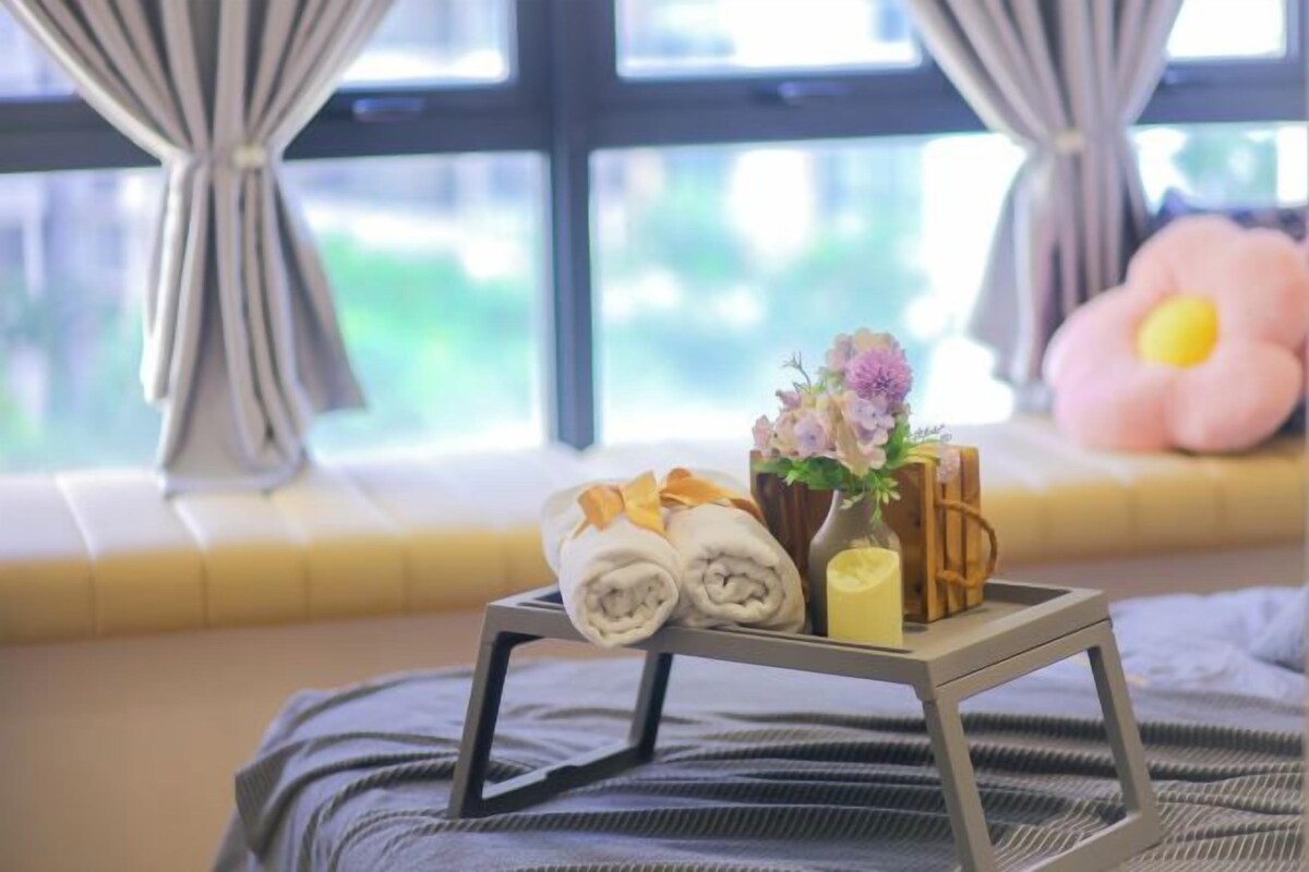 A neatly arranged tray with towels, a candle, and a flower vase on a bedside table by a window with curtains.