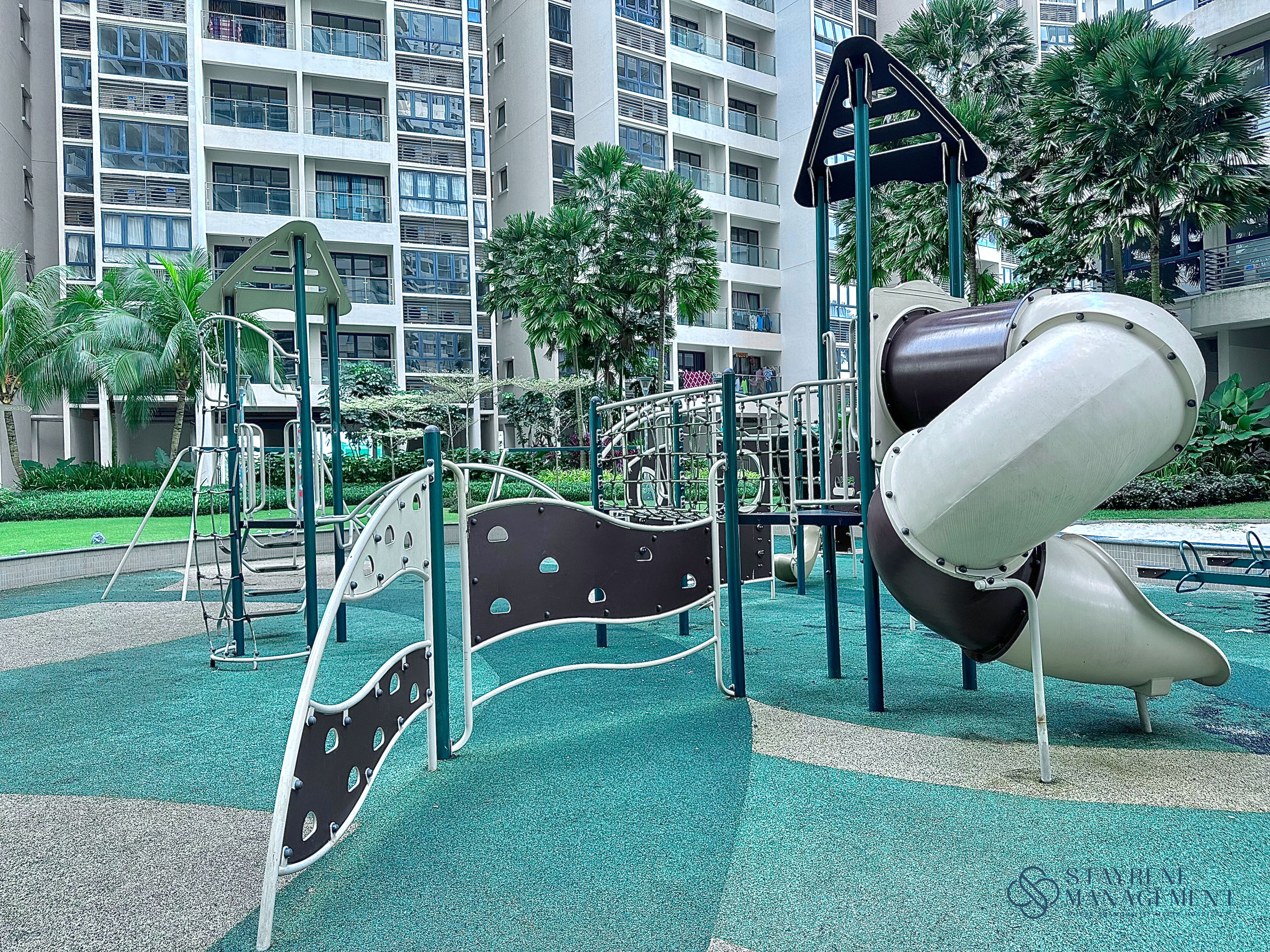 A playground with slides and climbing structures, surrounded by greenery and a multi-story building in the background.