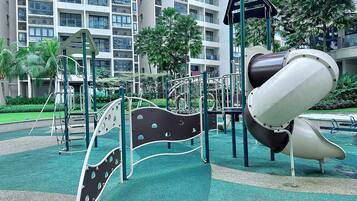 A playground with slides and climbing structures, surrounded by greenery and a multi-story building in the background.