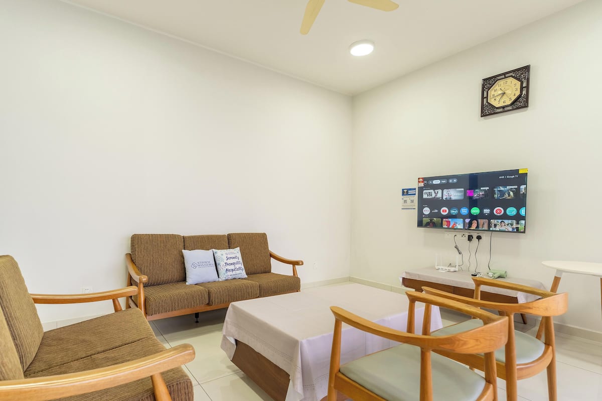 A room with a sofa, two wooden chairs, a small table, a TV mounted on the wall, and a clock.