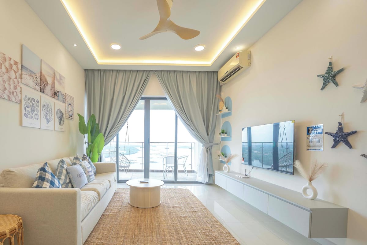 A modern living room with a sofa, a coffee table, and a TV. There is a large window with curtains, a ceiling fan, and wall-mounted air conditioning.