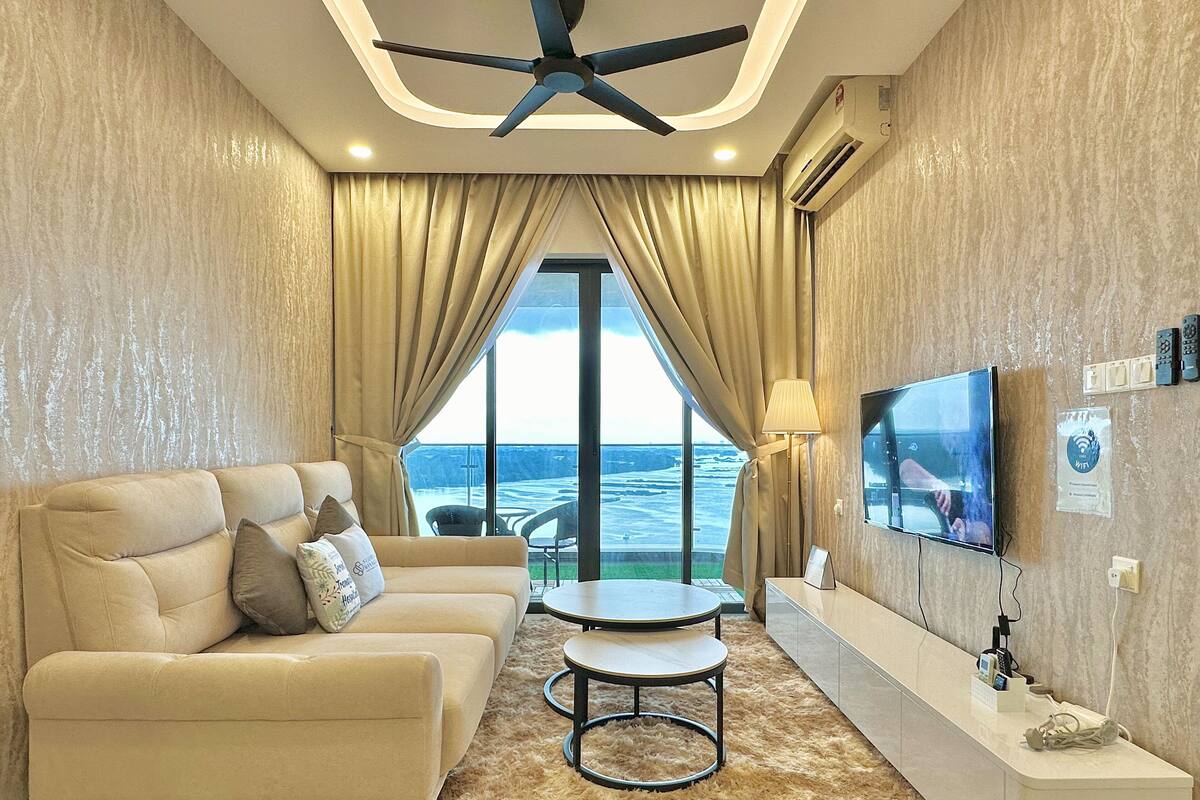 A modern living room with a sofa, a small table, a TV, and a view of the sea through large windows.