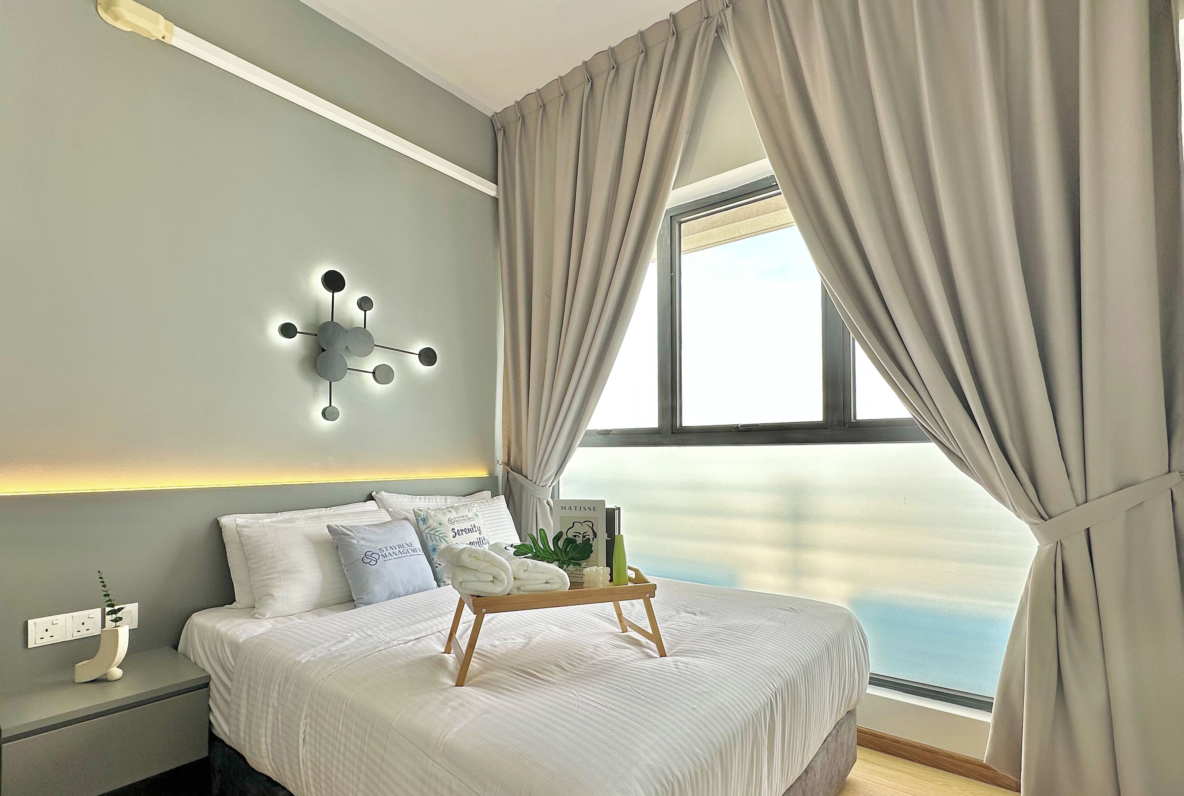 A neatly made bed with a tray of towels and toiletries, a modern wall light, and a view of the sea through a window with curtains.