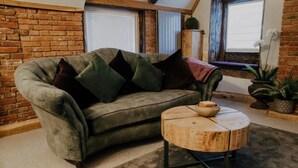 A cozy living room with a green sofa, multiple cushions, a round wooden coffee table, and a brick wall. - Urban Lofts, Melksham - Butler (Melksham)