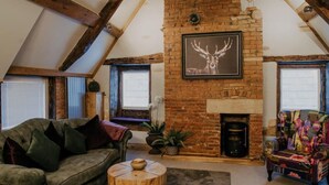 A cozy loft-style living room with exposed wooden beams, a brick fireplace, and a large framed deer head artwork. - Urban Lofts, Melksham - Butler (Melksham)