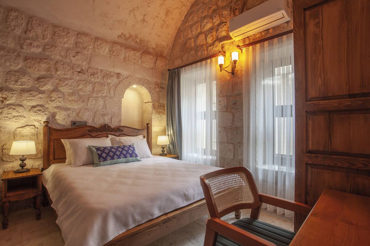 Un dormitorio con pared de piedra, cama de madera, mesita de noche con lámpara y ventana con cortinas.