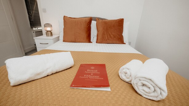 Cama bem arrumada com um livreto vermelho, dois travesseiros marrons e duas toalhas brancas enroladas.