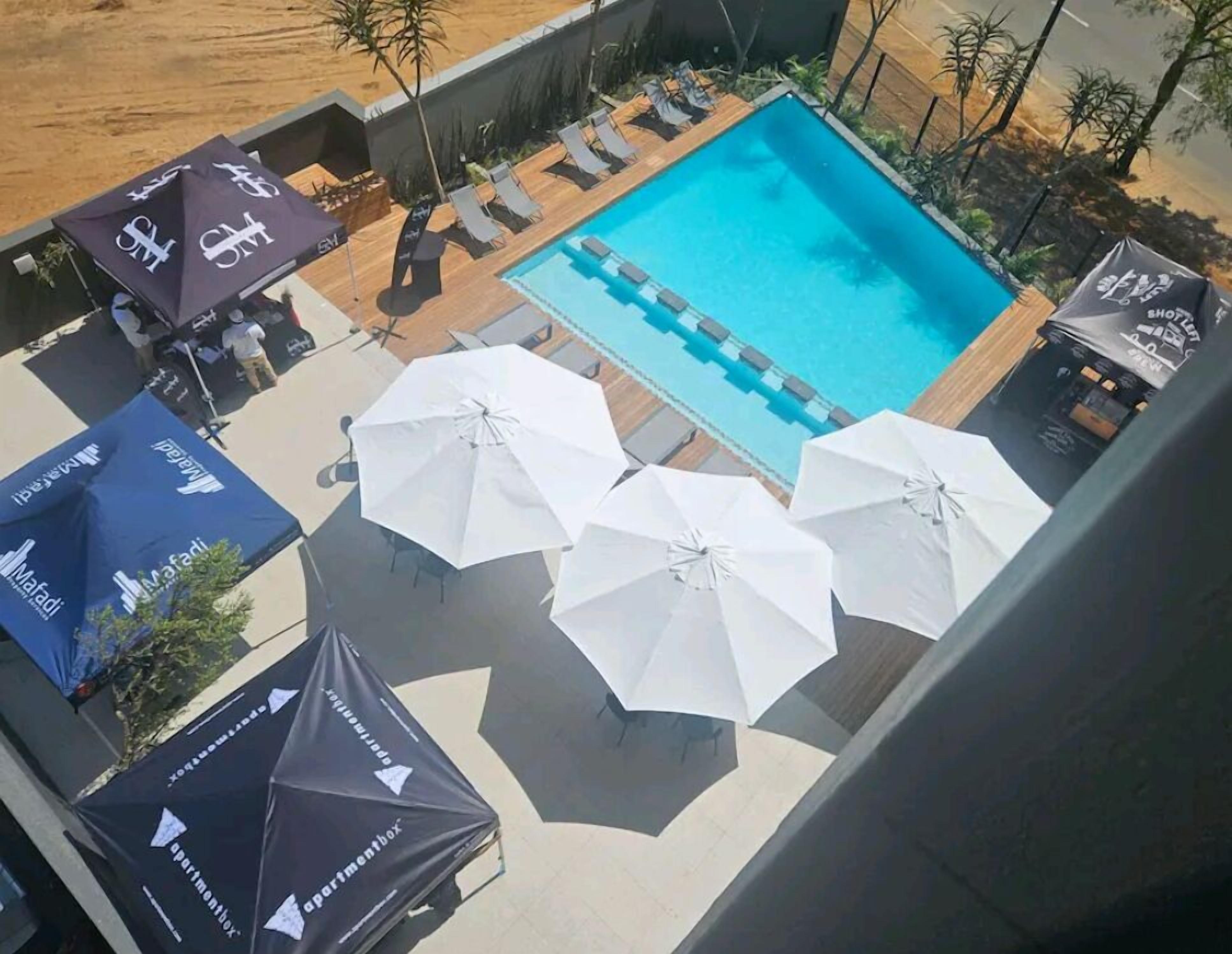 A rooftop pool area with lounge chairs and umbrellas, surrounded by a wooden deck and plants.