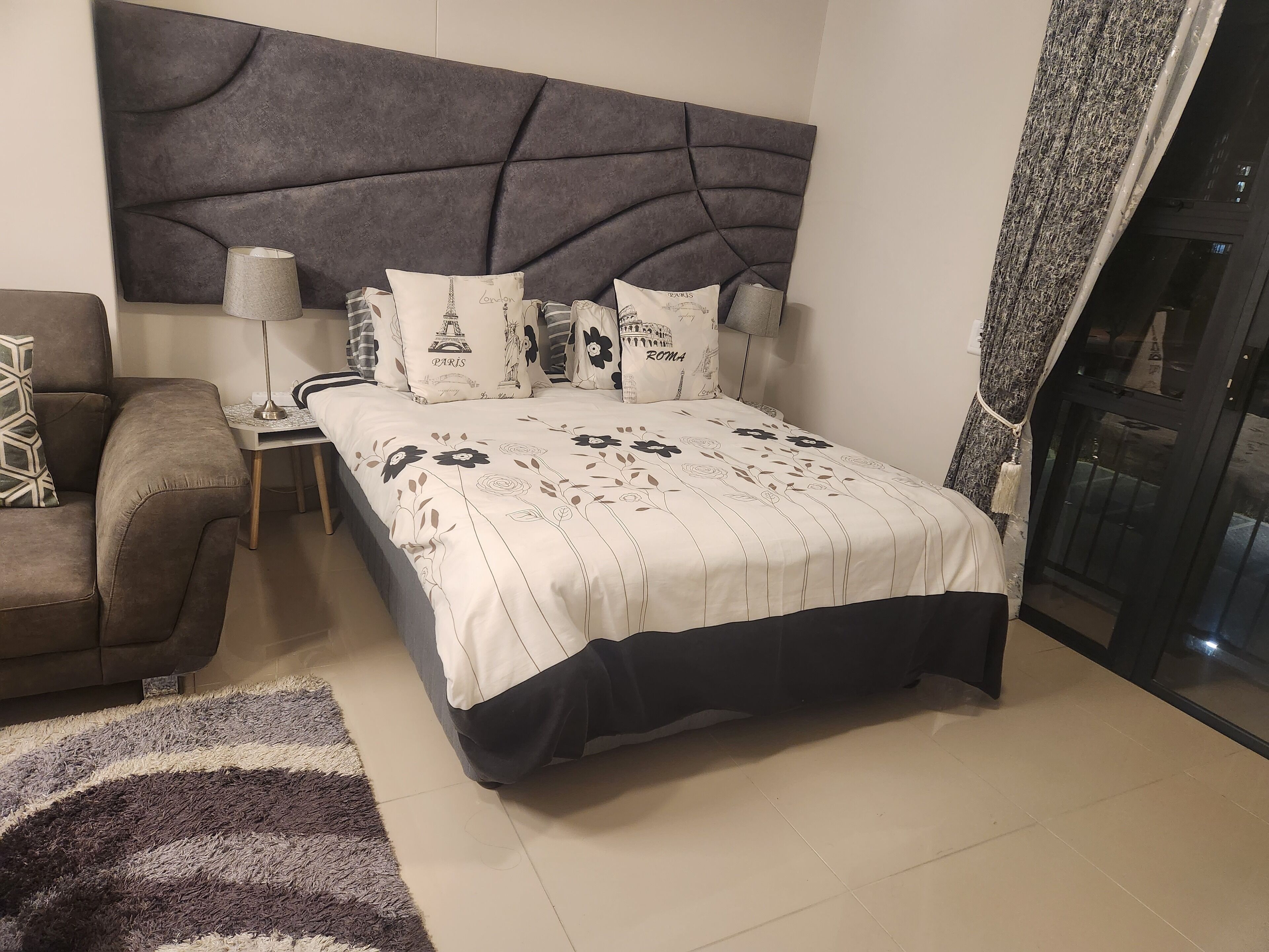 A modern bedroom with a large bed, a grey upholstered headboard, and decorative pillows with city names.