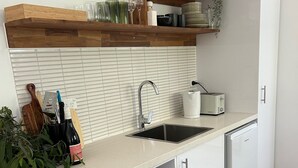 A modern kitchen with white cabinetry, a built-in microwave, a sink, and a toaster. There are wooden shelves above the counter holding glassware and a potted plant. -  Tangara - A Stylish Newly Renovated Studio Apartment, close to the surf beach. (Inverloch)