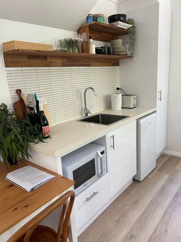 A modern kitchen with white cabinetry, a built-in microwave, a sink, and a toaster. There are wooden shelves above the counter holding glassware and a potted plant.