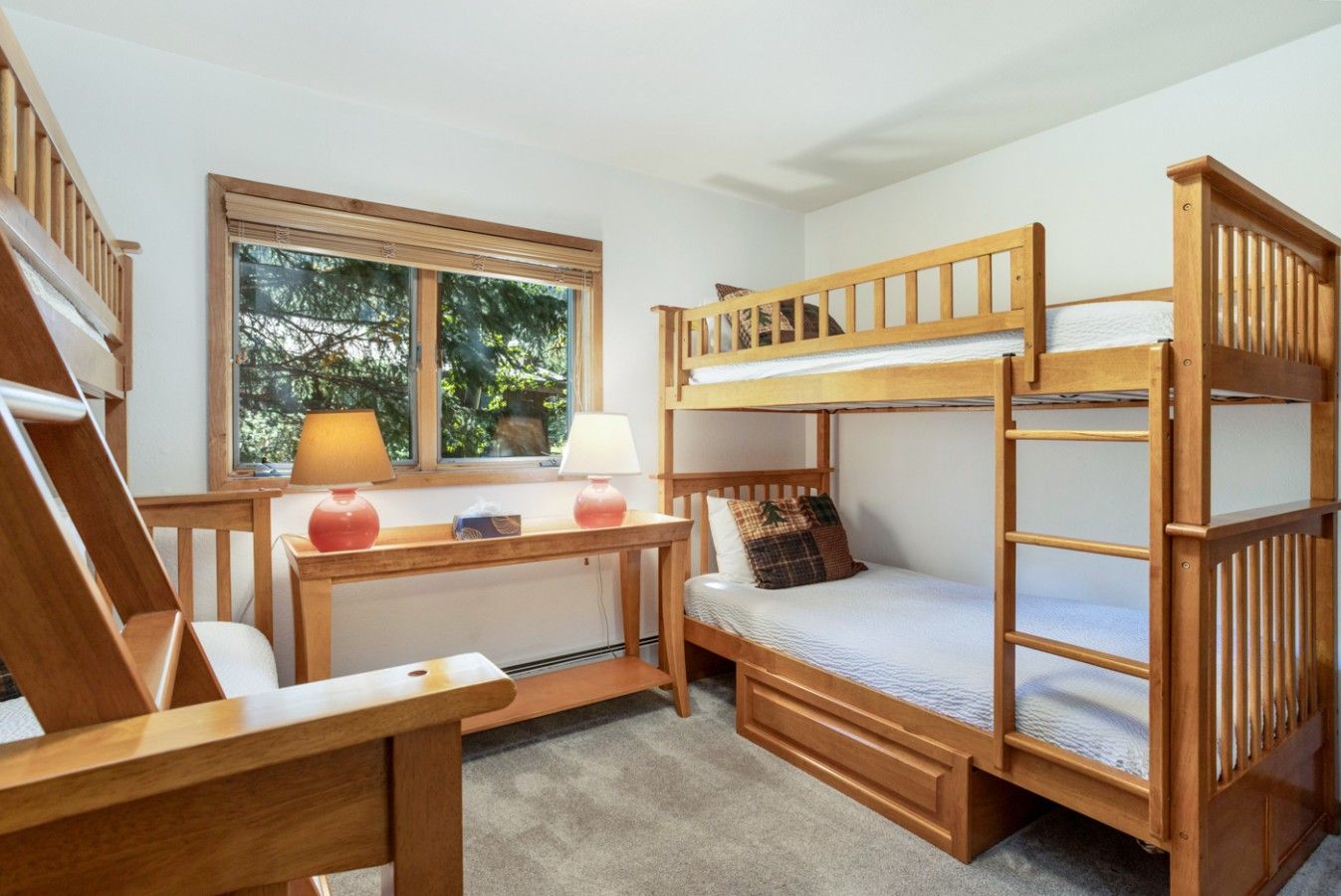 A bunk bed room with a desk and two lamps, a window with a view of trees, and a staircase.