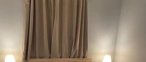 En snyggt bäddad säng med en hopvikt filt, två sänglampor, en sänggavel med geometriska kuddar och en gardin.