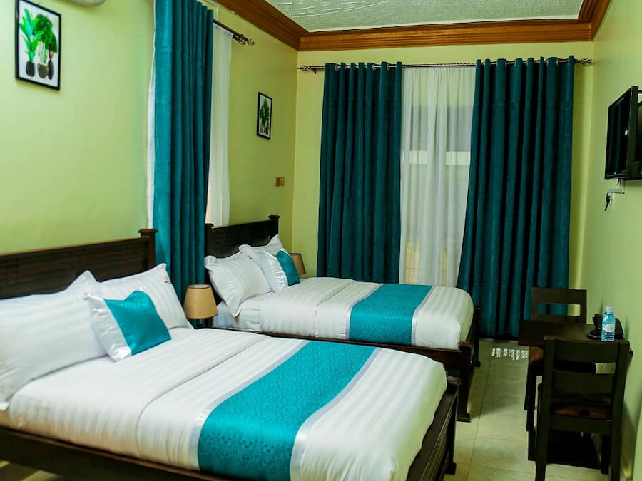 A hotel room with two beds, teal and white bedding, a wooden headboard, a desk with a chair, a television, and a window with blue curtains.