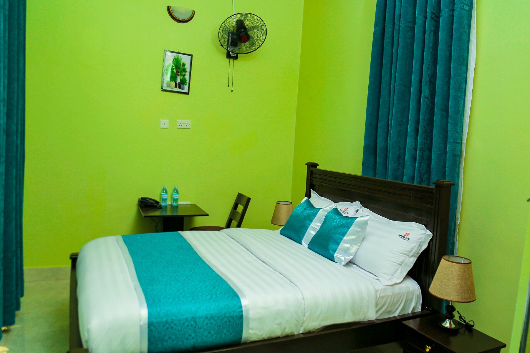 A hotel room with a bed, a small table with a lamp, a chair, and a wall-mounted fan.