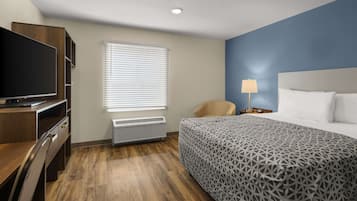 A hotel room with a bed, a desk with a TV, a chair, a lamp, and a window with blinds.