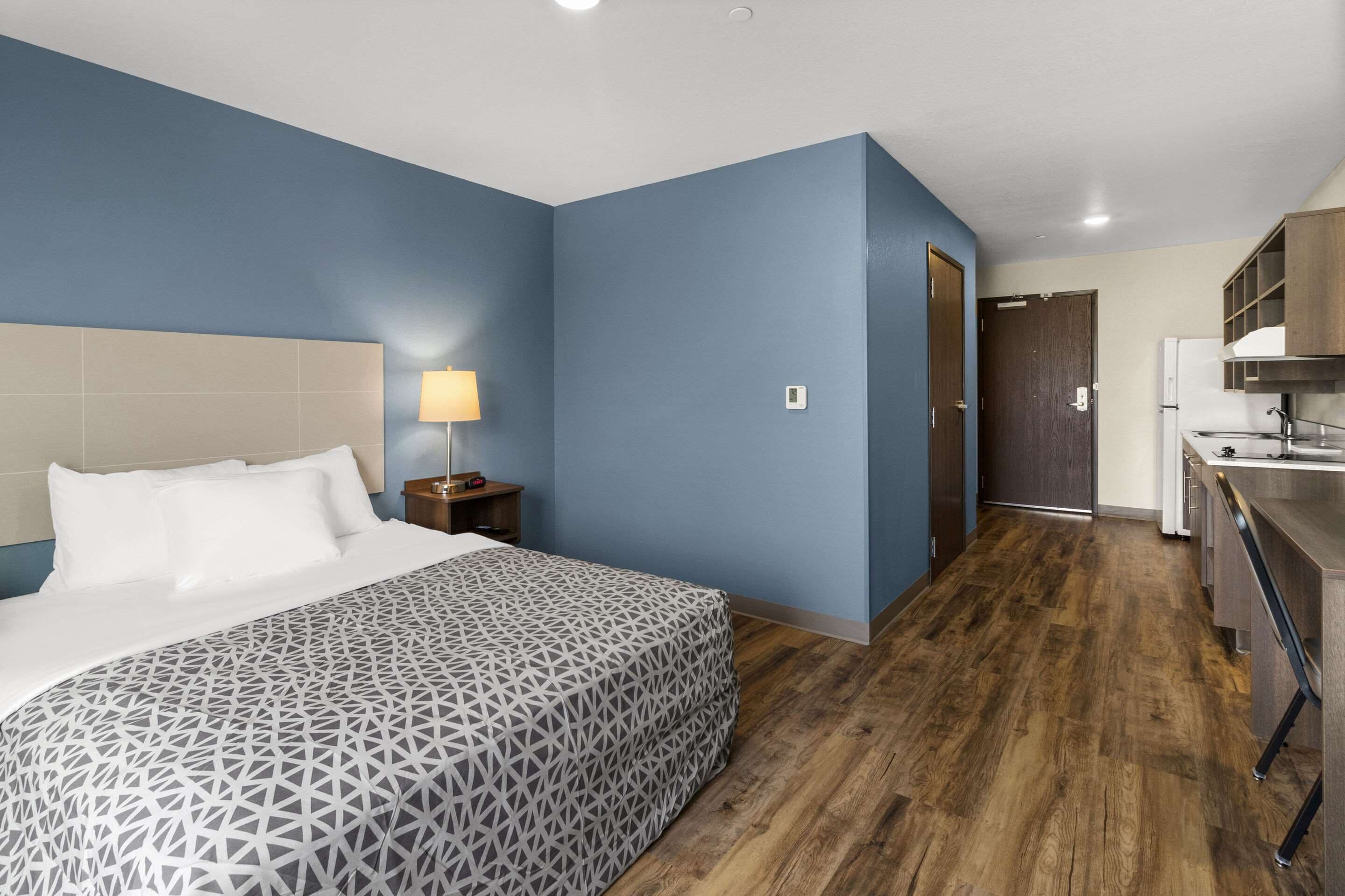 Una habitación de hotel con cama, mesita de noche, lámpara y una zona de kitchenette con refrigerador y lavabo.