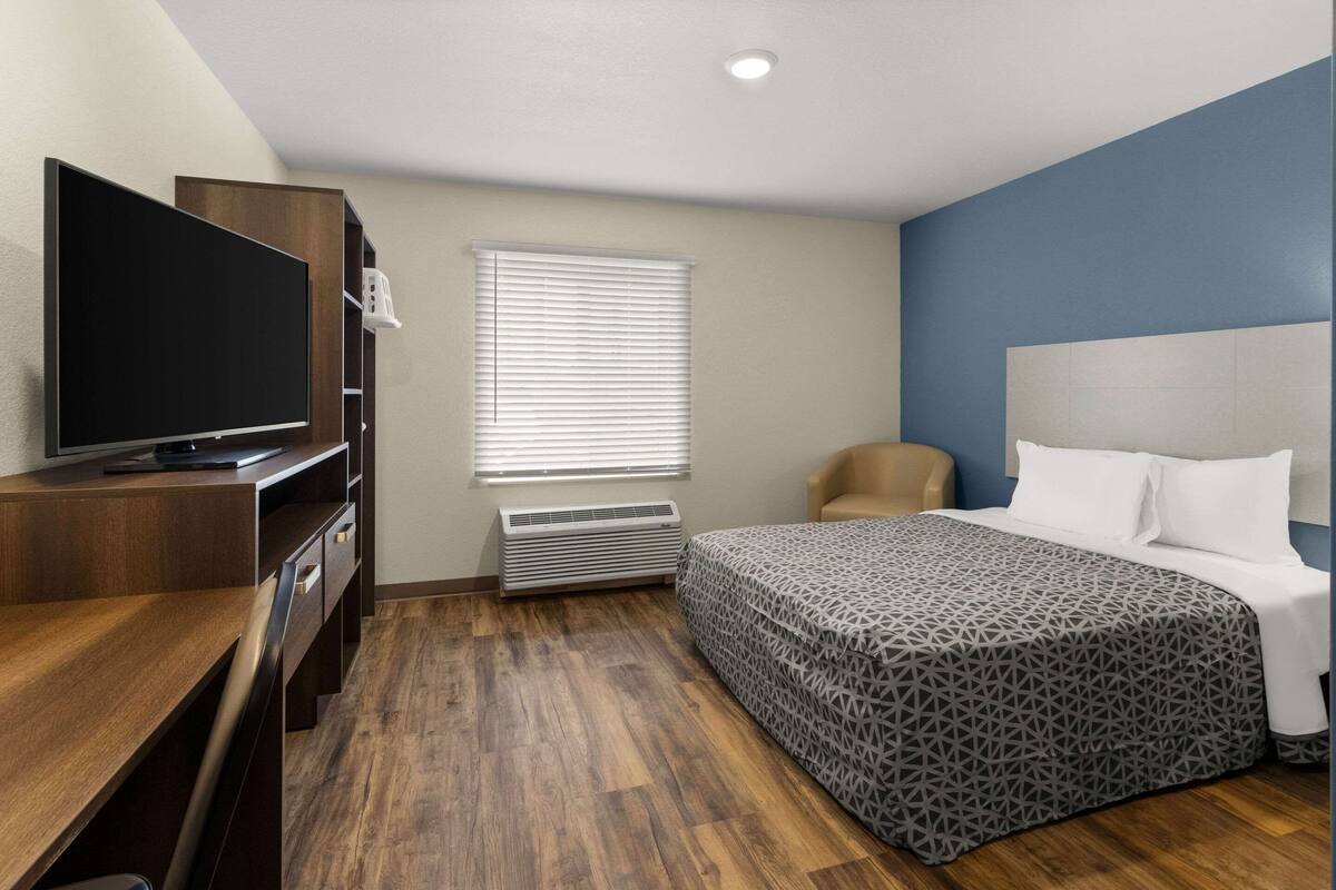 Habitación de hotel con un escritorio de madera, un televisor de pantalla plana, una cama con colcha estampada, una silla y una ventana con persianas.
