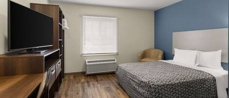 A hotel room with a wooden desk, a flat-screen TV, a bed with a patterned bedspread, a chair, and a window with blinds.