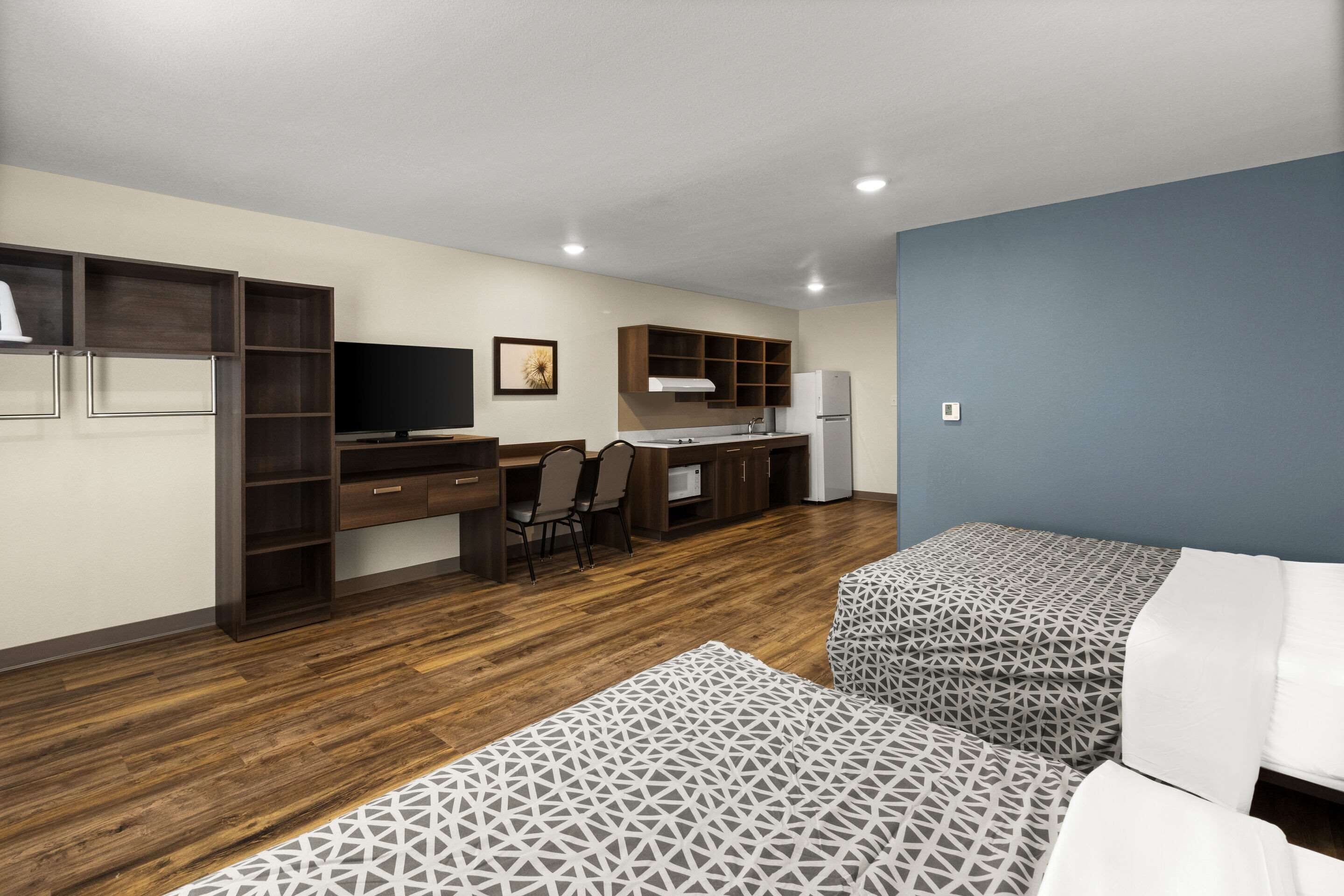 A compact room with a kitchenette, a desk, two beds with patterned bedding, and a television.