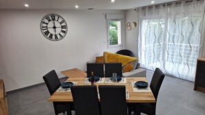 Un comedor con una mesa de madera dispuesta para cuatro, sillas negras, un reloj de pared grande y una ventana con cortinas translúcidas.