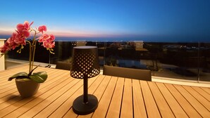 A wooden deck with a potted orchid and a perforated lamp on a balcony overlooking a cityscape at dusk. - Luxurous appartment with stunning views of the golfcourse and sea  (Orihuela Costa)