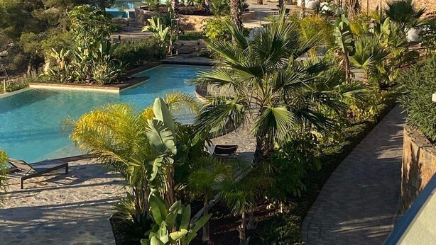 A resort with a swimming pool surrounded by lush greenery and palm trees, overlooking a mountainous landscape. - Luxurous appartment with stunning views of the golfcourse and sea  (Orihuela Costa)