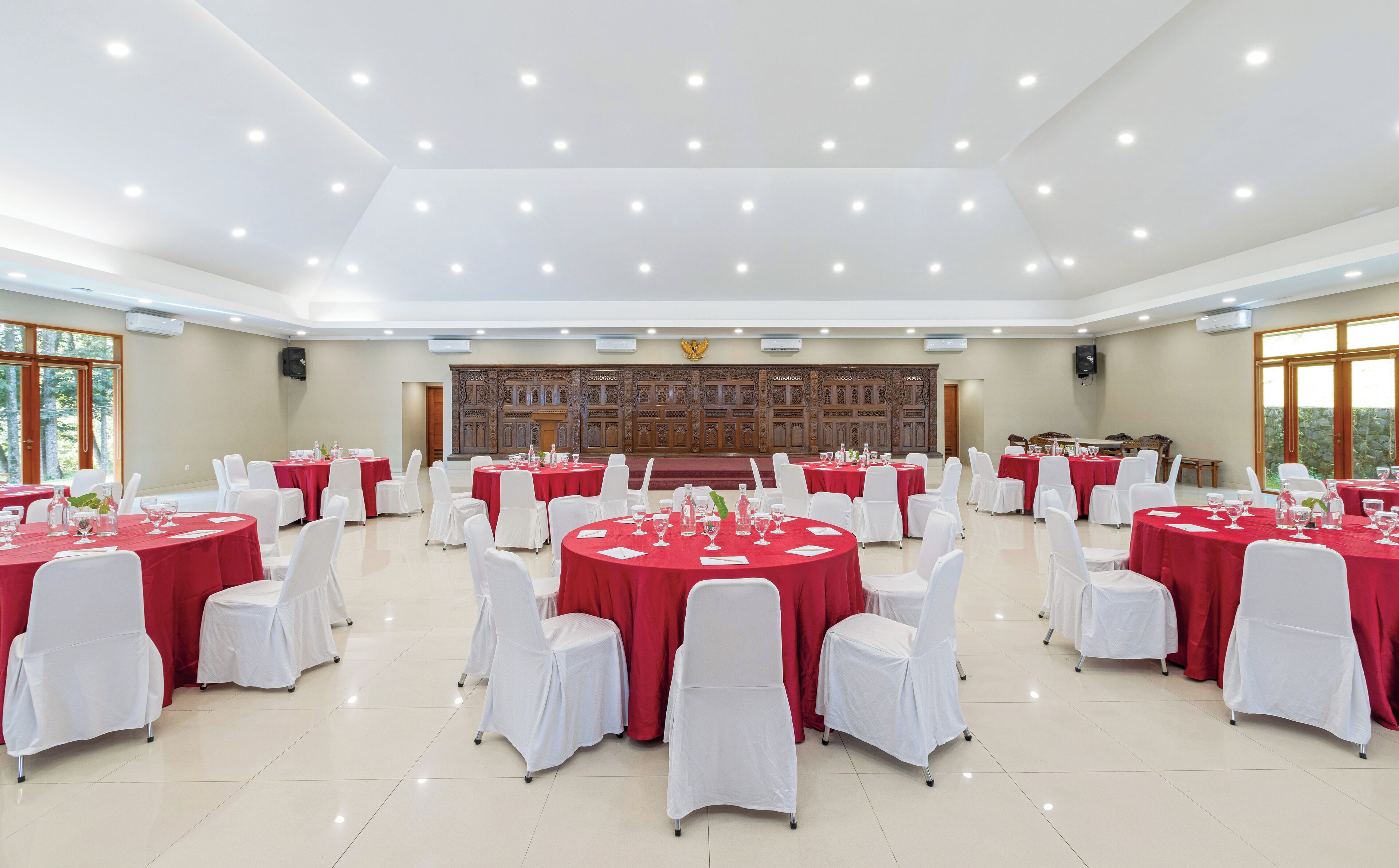 Un amplio salón de banquetes con mesas redondas cubiertas con manteles rojos y sillas blancas, un escenario con fondo de madera y grandes ventanales a la izquierda.