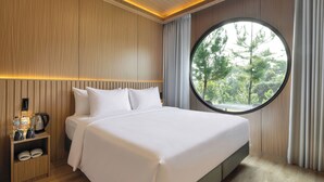 A modern hotel room with a large bed, a round window, and a nightstand with a kettle and coffee maker. - The Glamping @ARTOTEL Pelangi Park (Bogor)