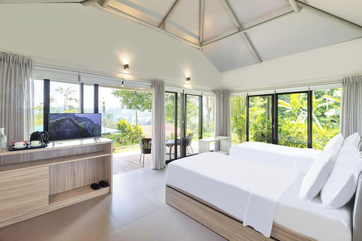 A modern hotel room with a large bed, a TV on a wooden console, a balcony with a table and chairs, and large windows with a view of greenery.